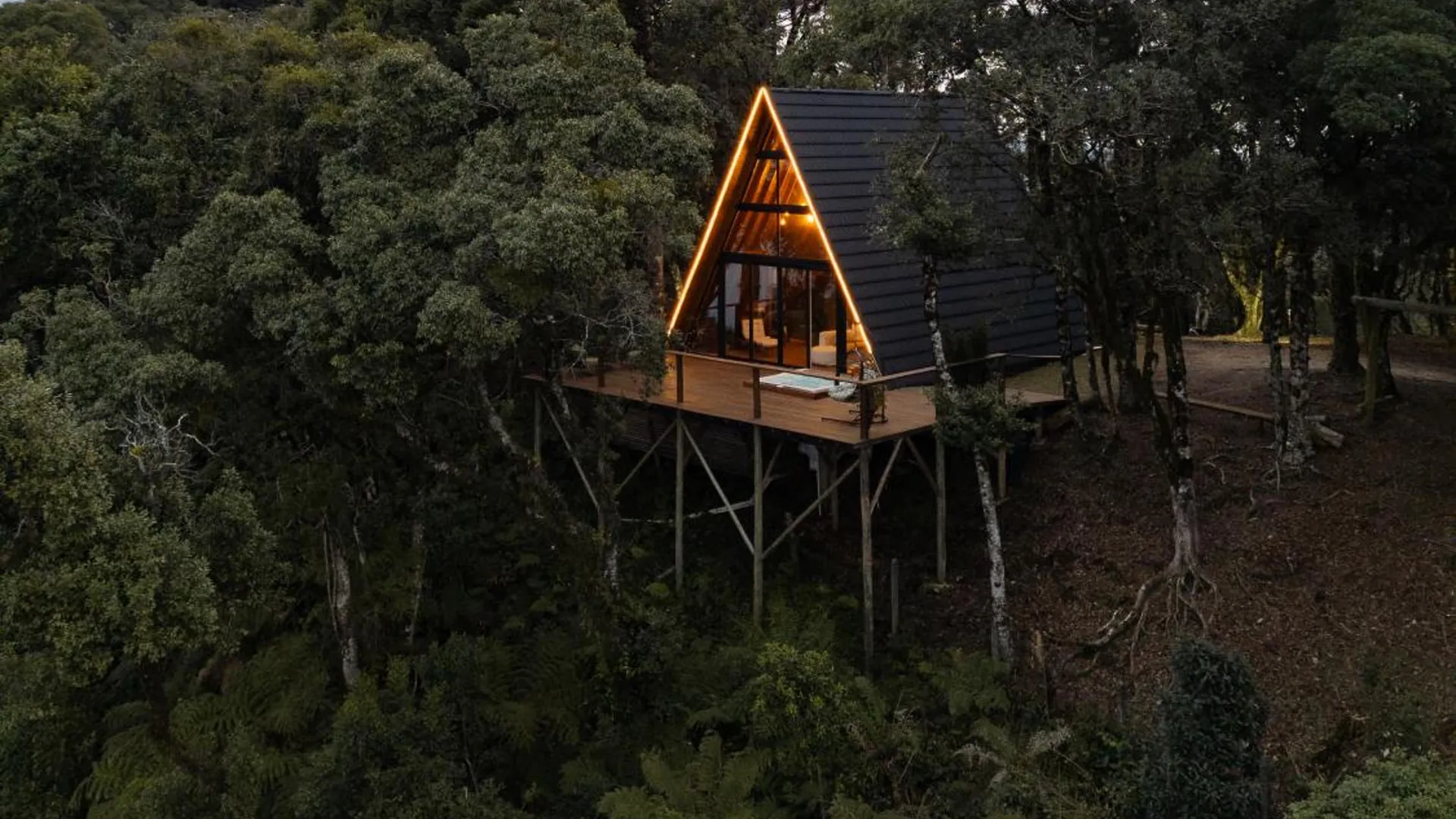 A Cabana Araucária acesa ao entardecer, sobre o deck suspenso entre as araucárias, vista do alto
